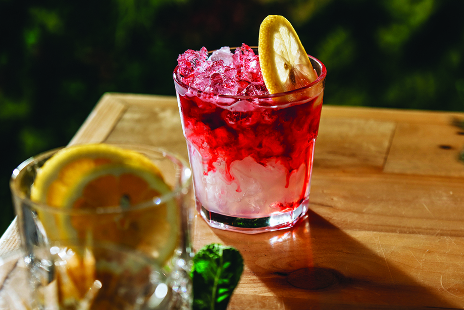 Cocktail with red colouring trickling down and a slice of lemon, with a glass full of lemons in foreground